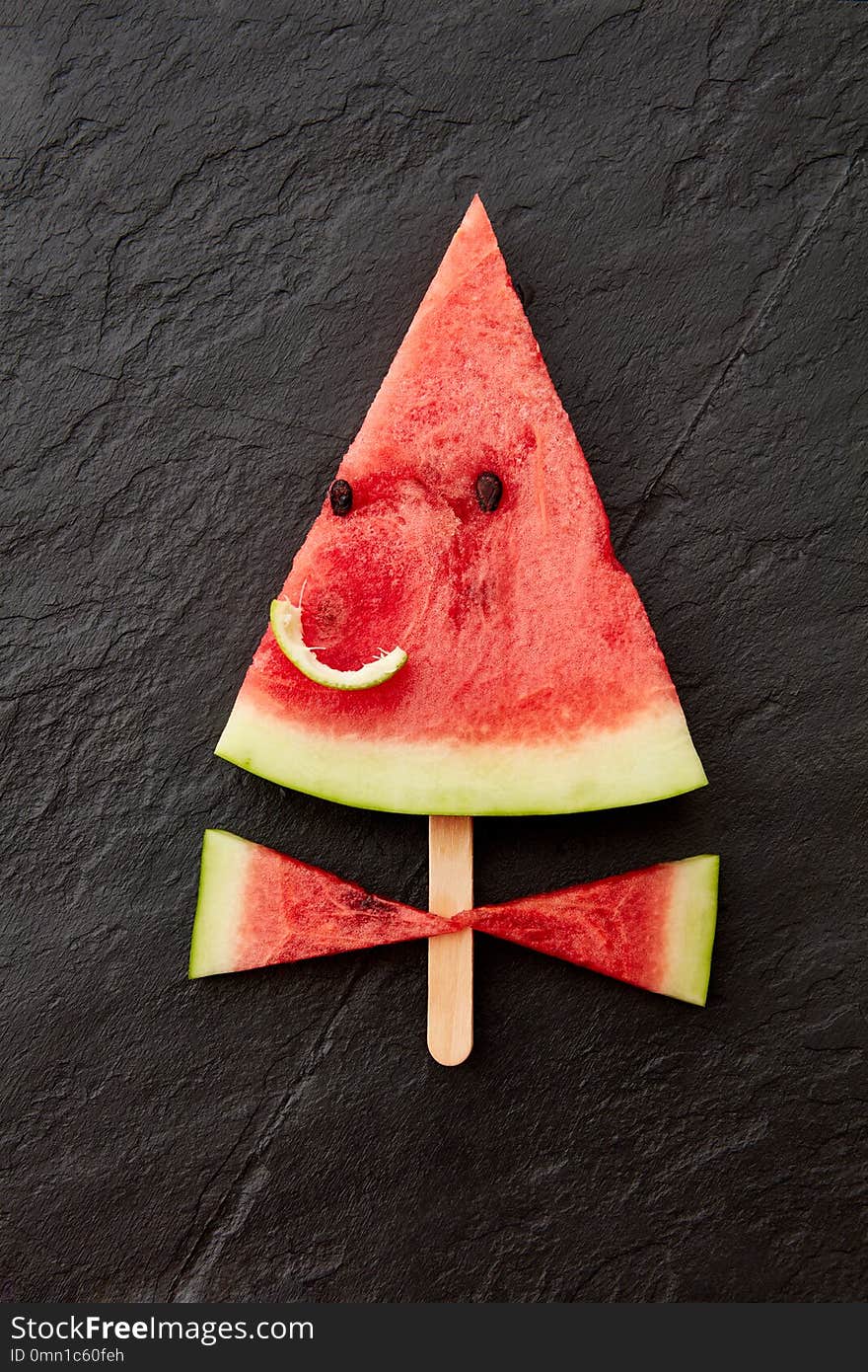 A juicy piece of watermelon on a stick in the form of a smiling face on a dark slate background with copy space. Flat