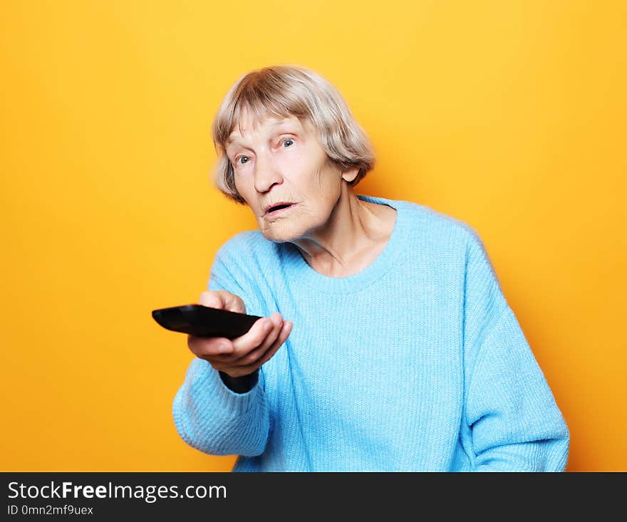 Lifestyle and people concept: funny grandma is holding a TV remote over yellow background, close up