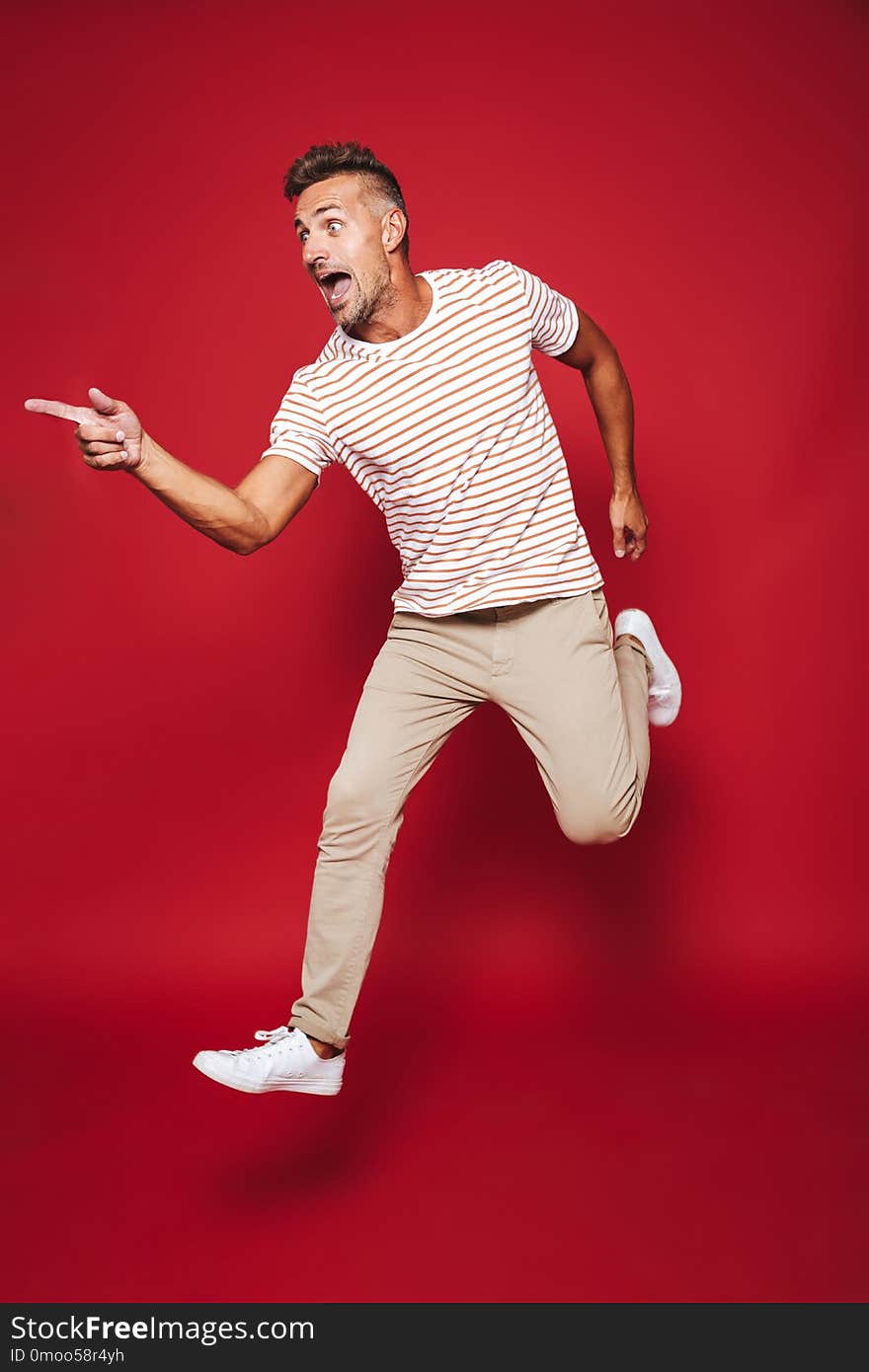 Full length photo of adult man in striped t-shirt running and screaming isolated over red background