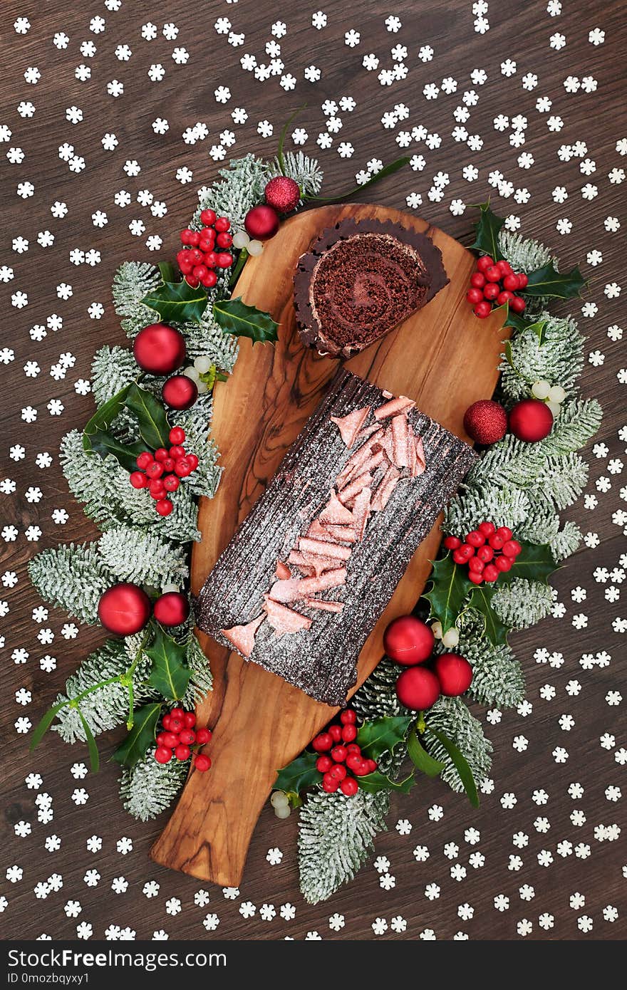 Chocolate log yule cake with winter flora of snow covered fir, holly and mistletoe on rustic wooden board and oak table background with snowflake sprinkles. Top view. Chocolate log yule cake with winter flora of snow covered fir, holly and mistletoe on rustic wooden board and oak table background with snowflake sprinkles. Top view.