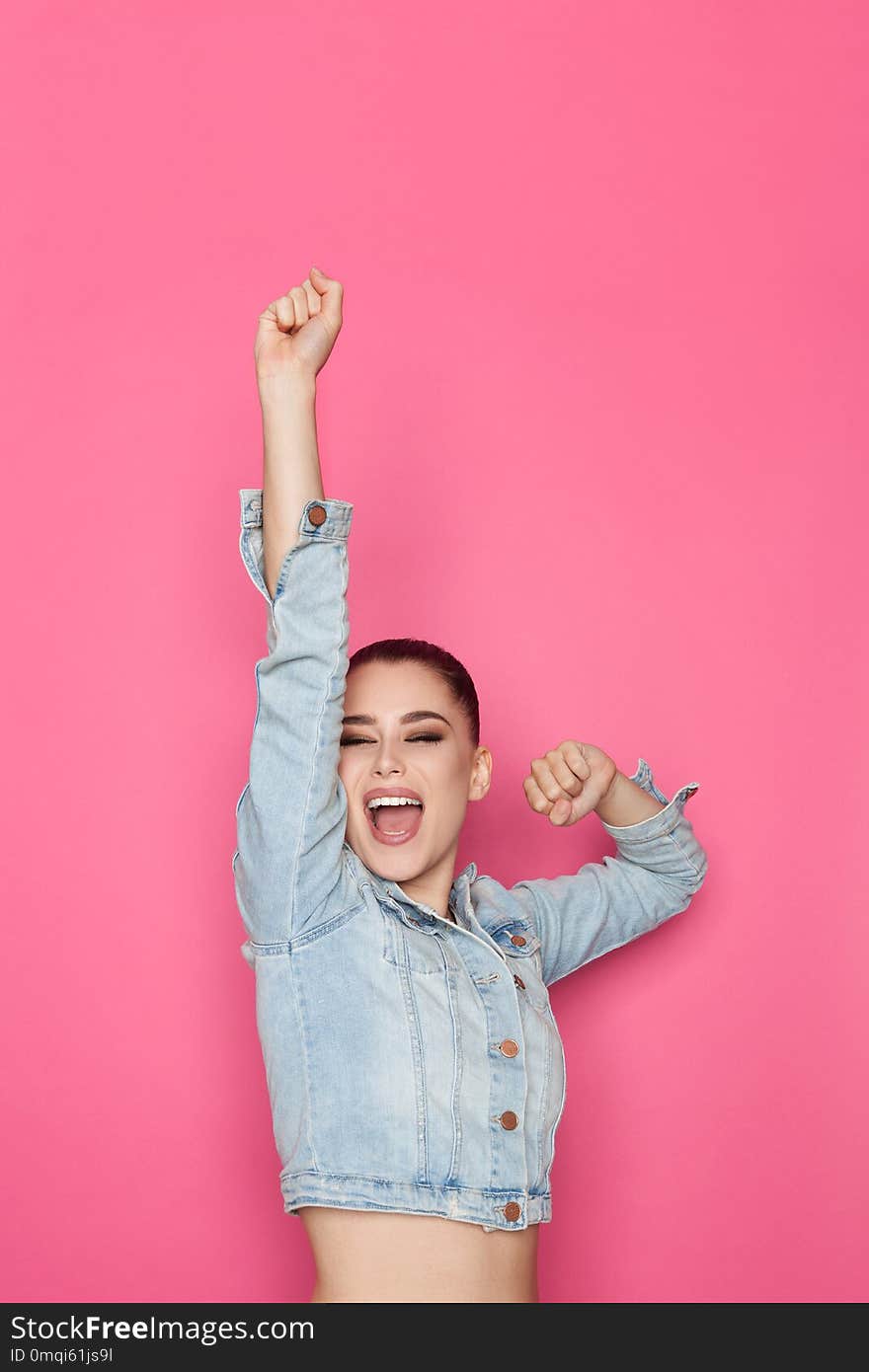 young woman in jeans jacket is holding arm raised and shouting. Waist up studio shot on pink background. young woman in jeans jacket is holding arm raised and shouting. Waist up studio shot on pink background.