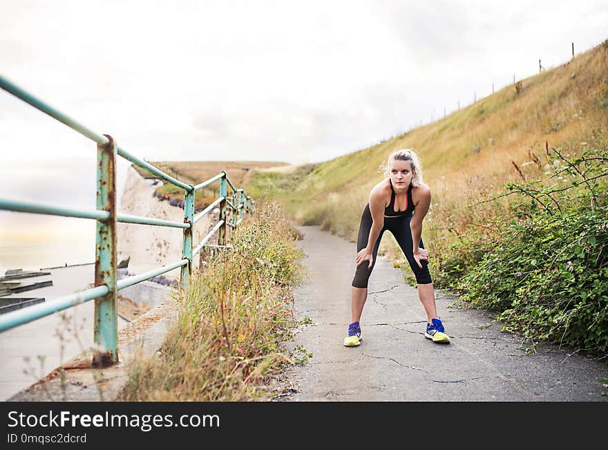 Young sporty woman runner in black activewear standing outside by the seaside, resting. Copy space.