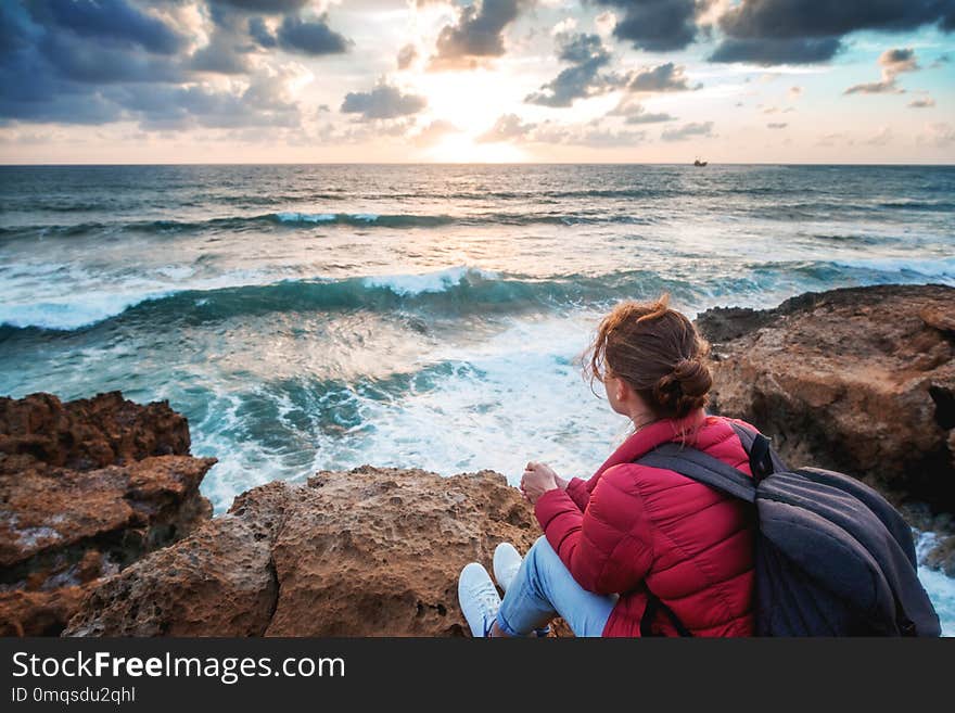 Young woman traveler sits on the shore of the winter sea and watches the bright colorful sunset