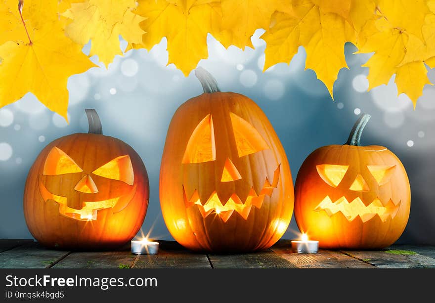 Three Halloween pumpkins head jack o lantern and candles on wooden table background , bokeh lights and autumn yellow maple leaves frame. Three Halloween pumpkins head jack o lantern and candles on wooden table background , bokeh lights and autumn yellow maple leaves frame