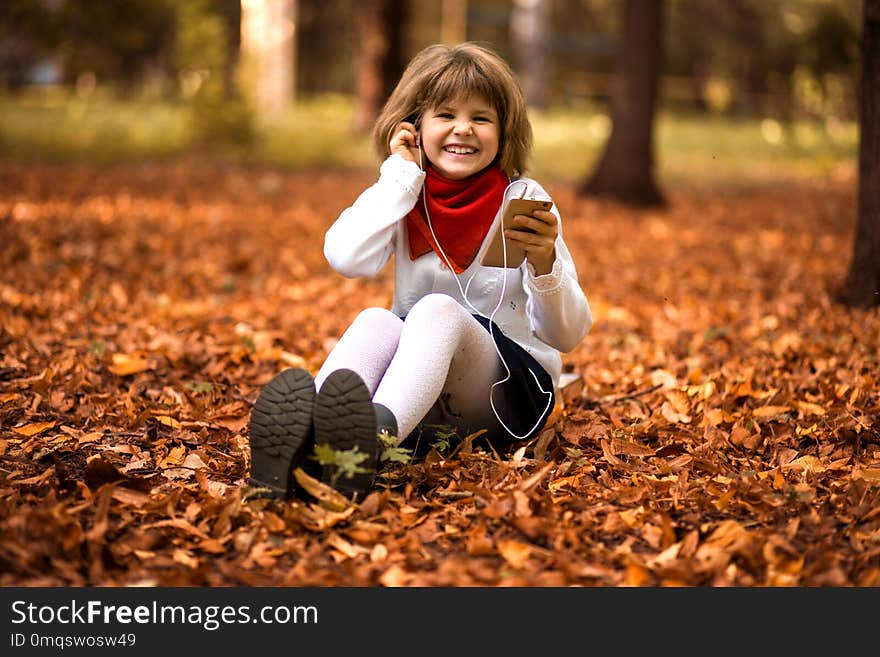 Happy little girl sits on yellow leaves and listens to music in autumn park