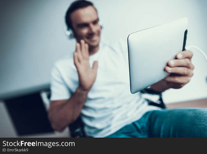 Blurred Disabled Man in Headphones Communicate Via Tablet. Smiling Person Wears T-Shirt and Blue Jeans Sits in Wheelchair Browsing Internet and Talking Via Video and Showing Hello Gesture. Blurred Disabled Man in Headphones Communicate Via Tablet. Smiling Person Wears T-Shirt and Blue Jeans Sits in Wheelchair Browsing Internet and Talking Via Video and Showing Hello Gesture.