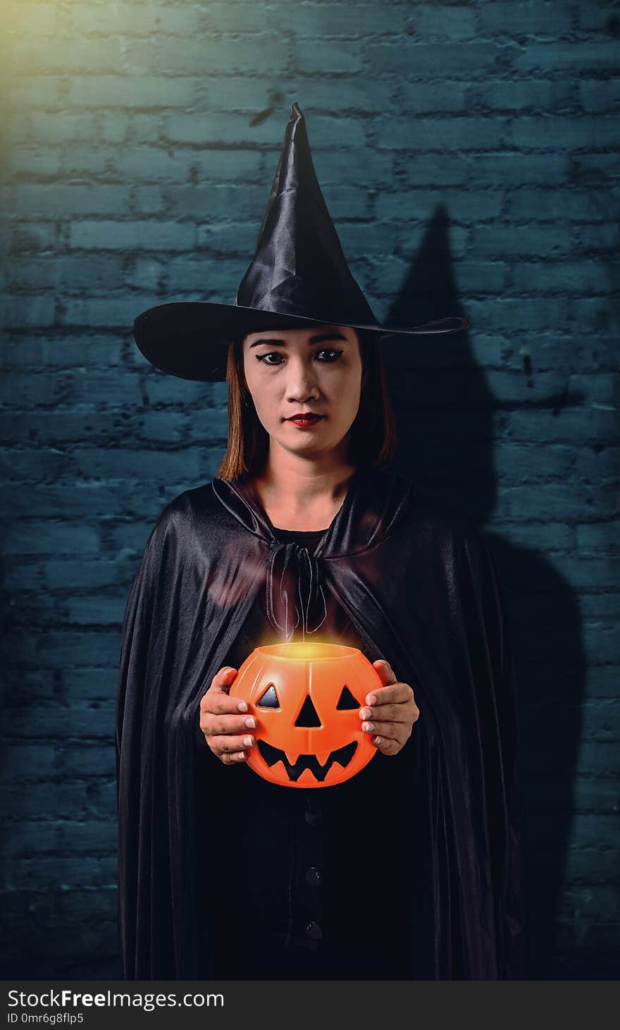 Portrait of woman in black Scary witch halloween costume holding halloween pumpkin with Old Brick Wall of the Ancient Castle of the Witch. Portrait of woman in black Scary witch halloween costume holding halloween pumpkin with Old Brick Wall of the Ancient Castle of the Witch