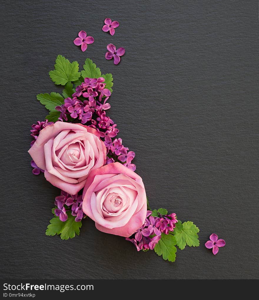 Decorative composition consisting of pink roses, violet lilac flowers and green leaves on a black slate with copy-space. Flat lay, top view, overhead view. Decorative composition consisting of pink roses, violet lilac flowers and green leaves on a black slate with copy-space. Flat lay, top view, overhead view