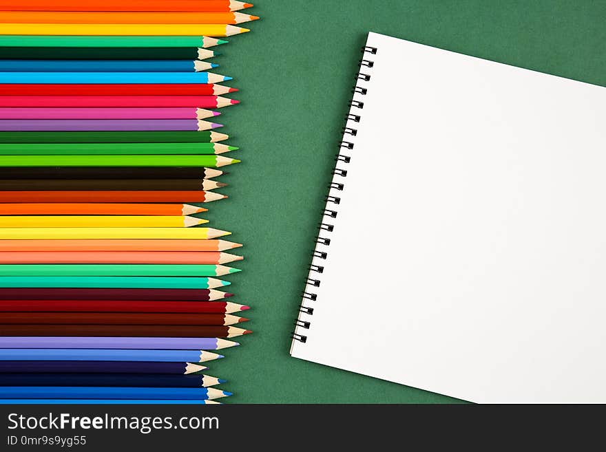 Top view of a set of many colorful pencils and empty sketchbook with a copy space on a green background. Art, craft, school and ed