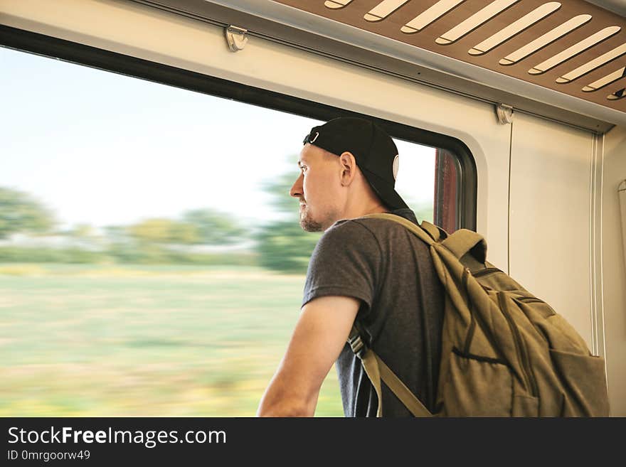 A young guy with a backpack inside the train looks out the window while the train is moving. A young guy with a backpack inside the train looks out the window while the train is moving.