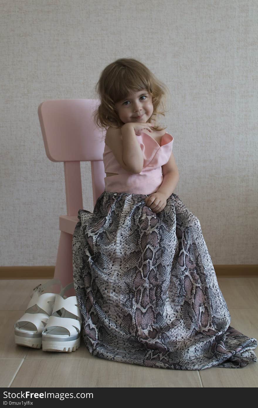 Little Funny Girl Sitting In A Chair In Her Mother`s Big Dress