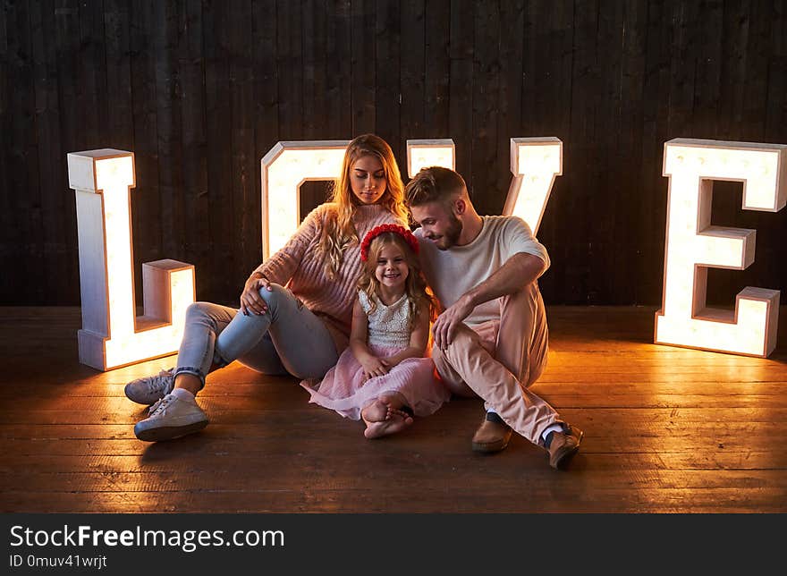 Mom, dad and daughter sits together in a room decorated with voluminous letters with illumination.