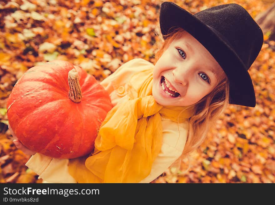 Little beautiful blond girl with big pumpkin in autumn background