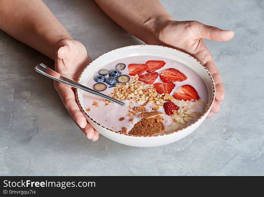 White bowl in a females hands with homemade granola, sliced strawberries, yogurt, blueberry, spoon - natural breakfast on a gray background. White bowl in a females hands with homemade granola, sliced strawberries, yogurt, blueberry, spoon - natural breakfast on a gray background.