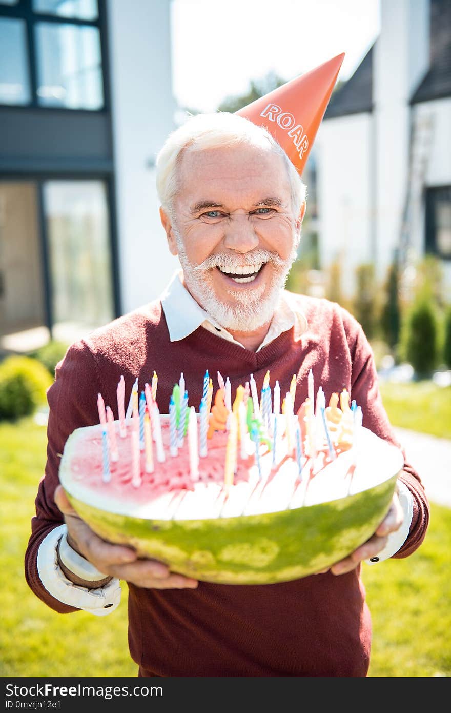 Funny celebration. Cheerful pensioner wearing paper hat while demonstrating his watermelon with candles. Funny celebration. Cheerful pensioner wearing paper hat while demonstrating his watermelon with candles