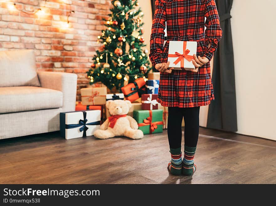 Little girl child holding gift in her back in the living room. attractive girl looking at the gifts and the teddy bear under christmas tree. cute kid with colorful socks standing on the wooden floor.