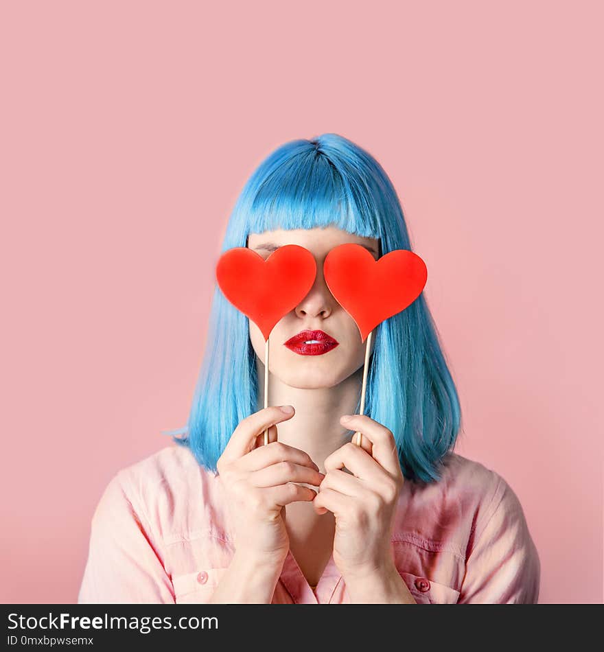 Young stylish woman with blue hairstyle and red lipstick holding valnetine`s heart on her eyes. Hipster funny girl model over pink pastel studio background. Love and valentine`s day design concept.