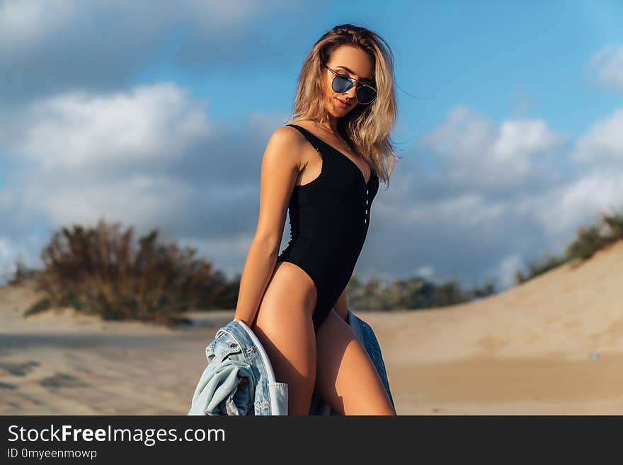 girl on the beach in a black swimsuit and blue sunglasses with a denim jacket in her hands. Beautiful model with a sports figure walks near the ocean on vacation, enjoying nature