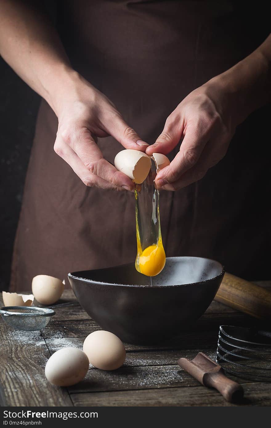 Professional chef hands are breaking an egg into bowl to make dough on wooden table, over dark background