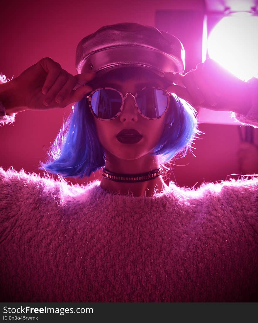 Young beautiful hipster woman in street style fashion winter concept wearing gold hat cap and round sunglasses in pink neon light. Young beautiful hipster woman in street style fashion winter concept wearing gold hat cap and round sunglasses in pink neon light