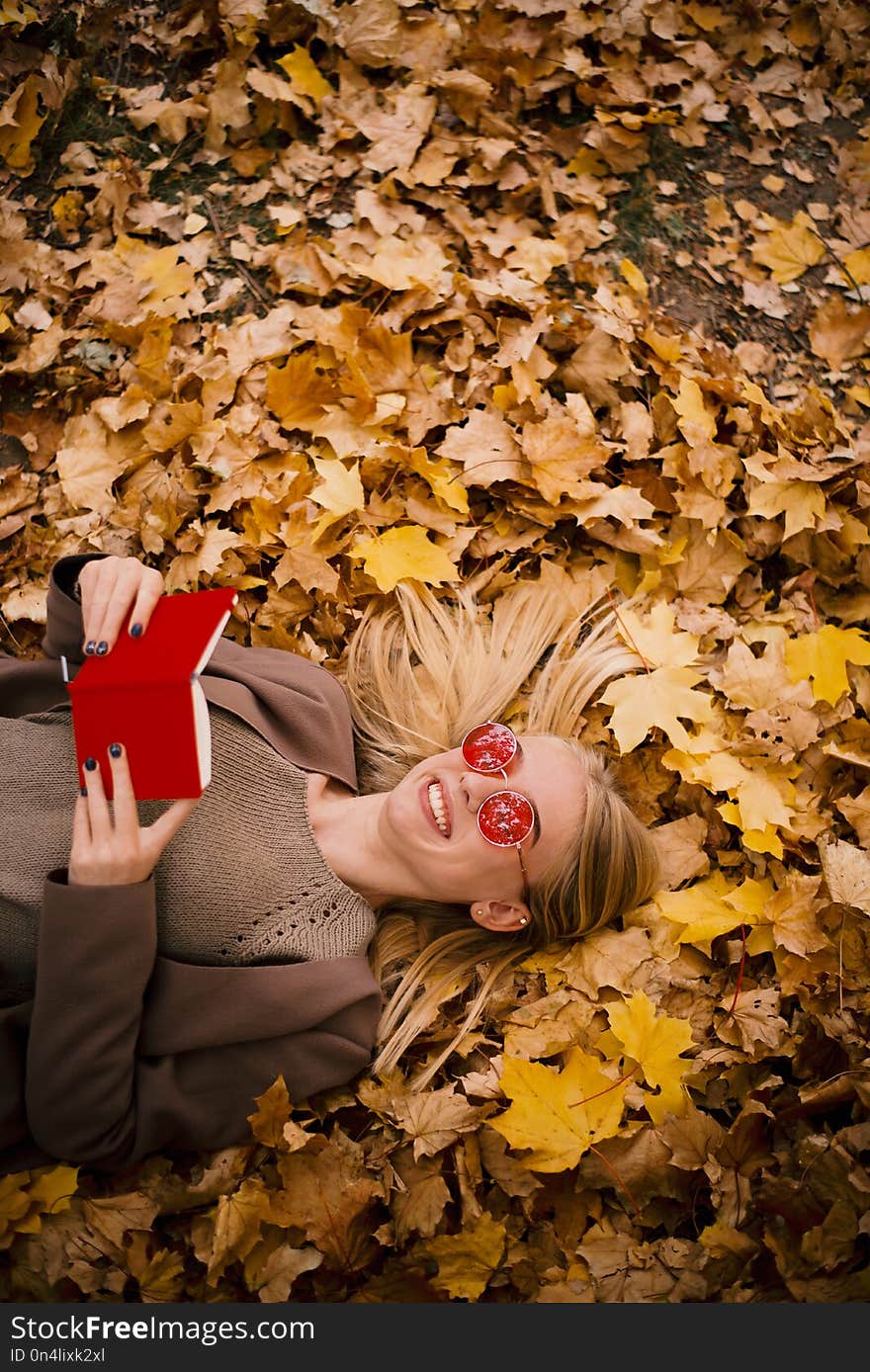 Beautiful young blonde in pink glasses lies in yellow aun leaves, reading a book in red cover, copyspace, vertical photo. Beautiful young blonde in pink glasses lies in yellow aun leaves, reading a book in red cover, copyspace, vertical photo