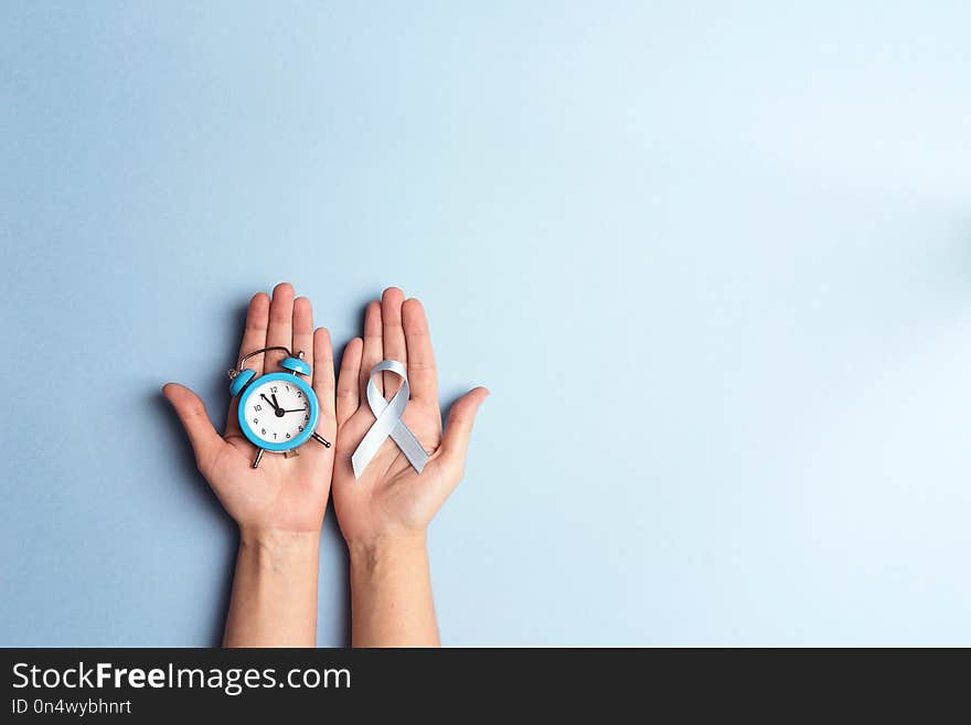 Helping hands holding blue ribbon and alarm clock on a blue background. World diabetes day,14 november. Copy space, top view.