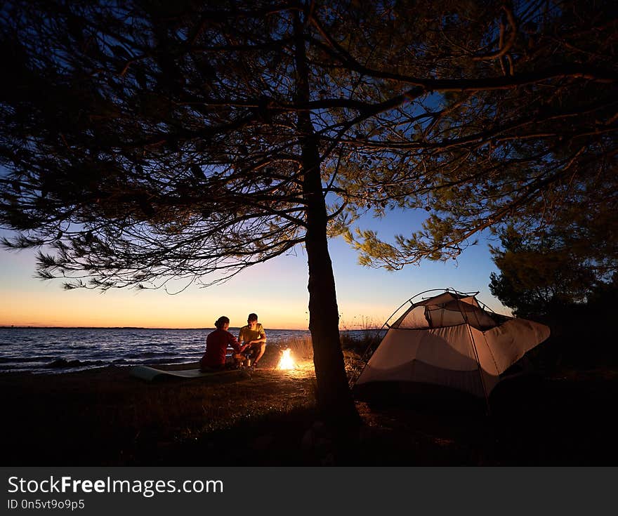 Young couple man and woman having rest at tourist tent and burning campfire on sea shore near forest