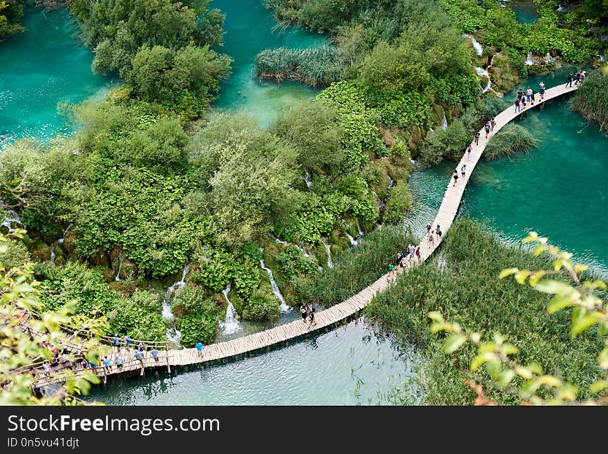 Plitvice Lakes National Park - UNESCO World Heritage Centre. Aerial view, colorful spring panorama of lit by sun green forest and long wooden bridge with tourists over blue lake with blue-green water.