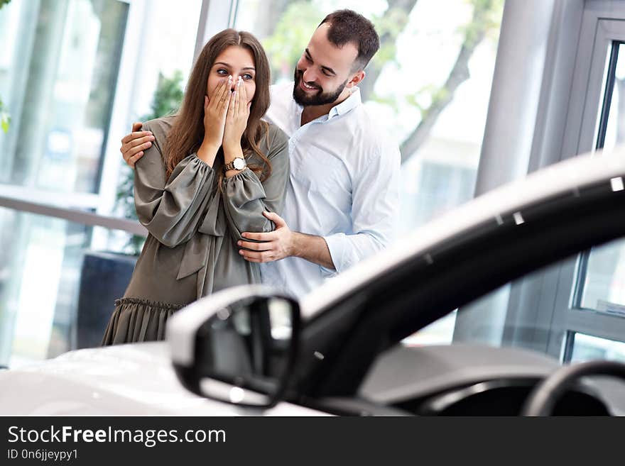 Adult man making surprise to beautiful woman in car showroom