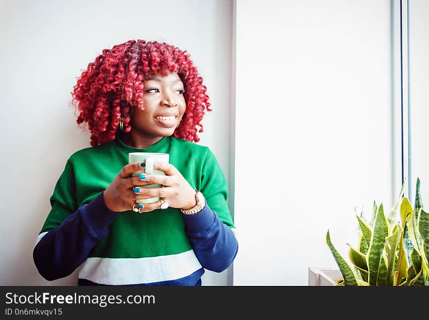 Young hipster dark-skinned woman dressed in casual clothes holding cup of hot drink, enjoying coffee or tea. Black student girl spending morning at cafe