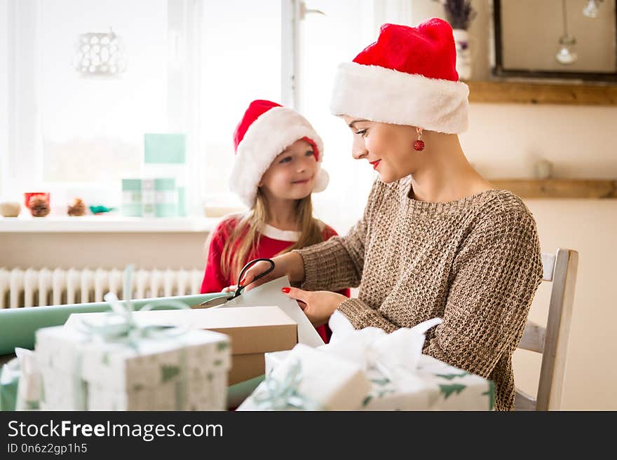 Mother and daughter wearing santa hats having fun wrapping christmas gifts together in living room. Candid family christmas time.