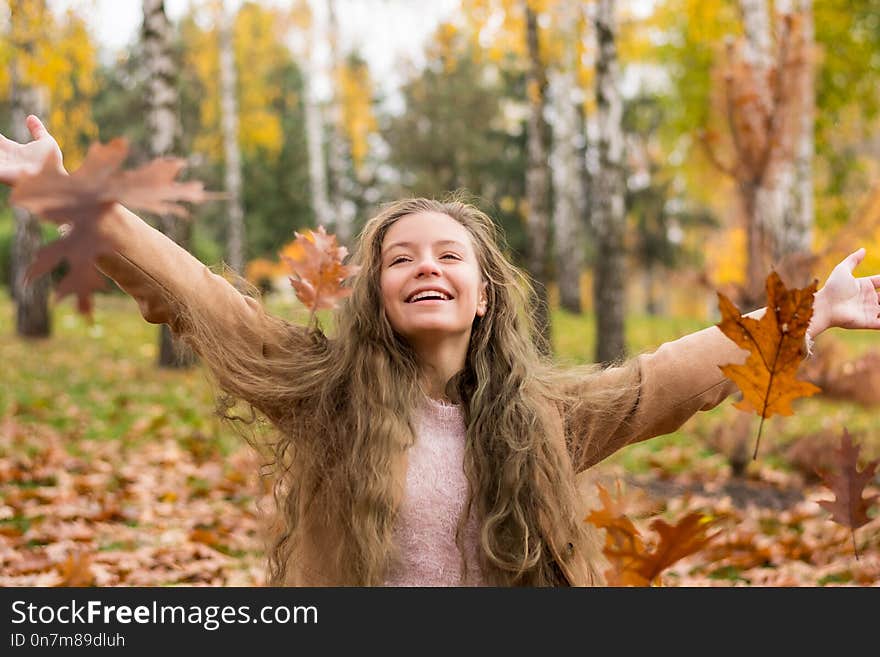 A teenager girl in a coat laughs and throws up leaves in autumn