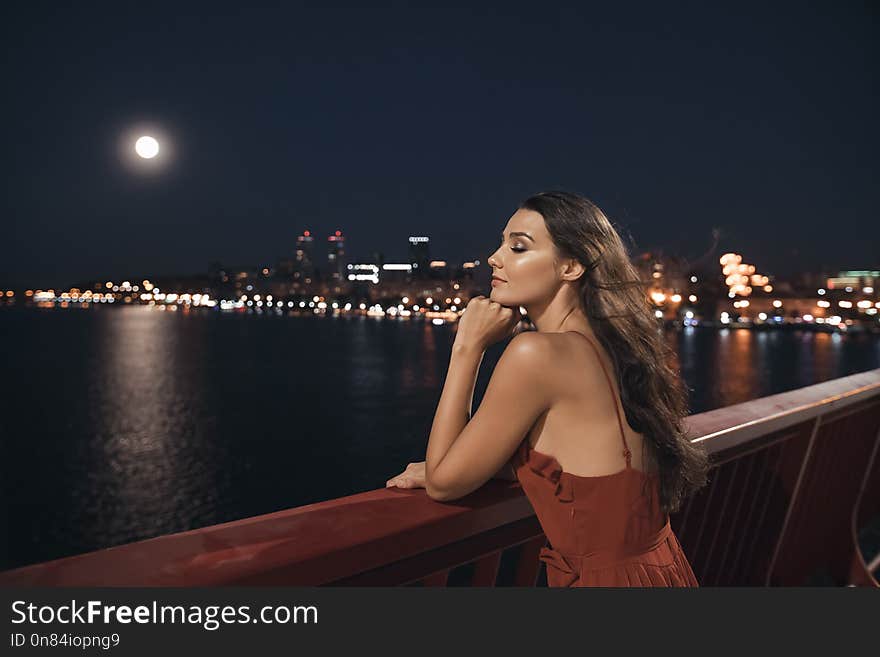 Young ellegant woman standing on the ligths of the night city background on bridge. romantic beautiful young mixed race Caucasian Asian girl with perfect makeup in long red evening dress standing over view on night river and moon in bokeh. Freedom, in love, dating concept. Young ellegant woman standing on the ligths of the night city background on bridge. romantic beautiful young mixed race Caucasian Asian girl with perfect makeup in long red evening dress standing over view on night river and moon in bokeh. Freedom, in love, dating concept