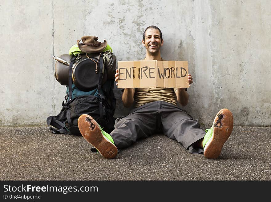 A single white adult man with a packed backpack sitting on the roadside in front of a gray wall wearing broken green shoes and holding a cardboard sign reading entire world. A single white adult man with a packed backpack sitting on the roadside in front of a gray wall wearing broken green shoes and holding a cardboard sign reading entire world