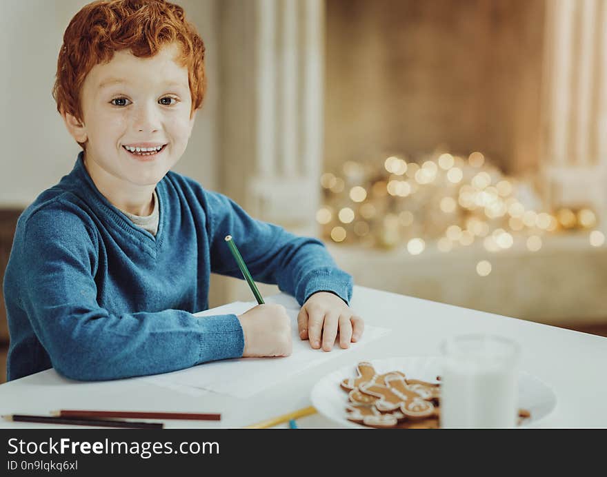 Feeling happiness. Cheerful kid keeping smile on his face while looking at camera while sitting at the table. Feeling happiness. Cheerful kid keeping smile on his face while looking at camera while sitting at the table