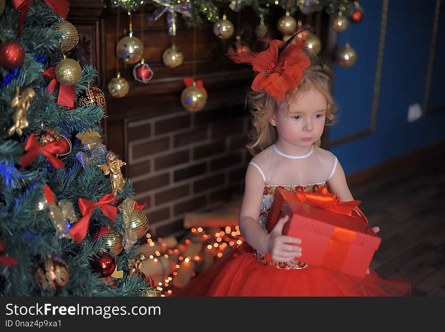 Cute little girl at the Christmas tree in a red elegant dress with a box with gifts in her hands
