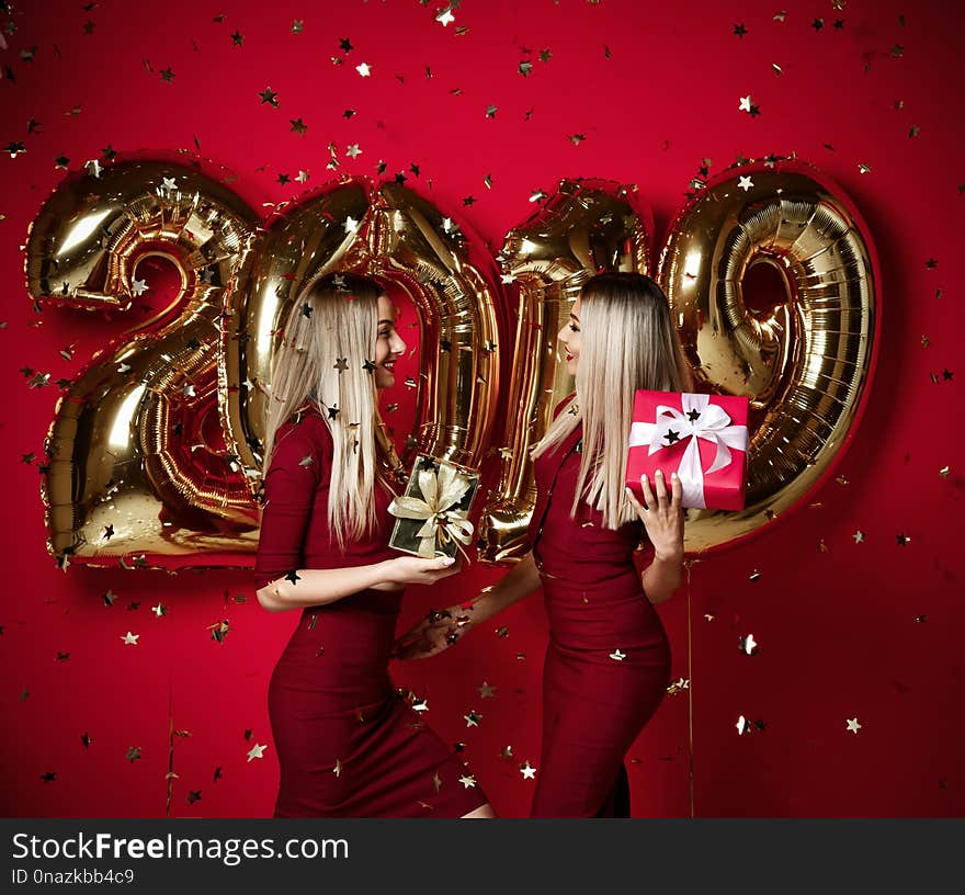 Two women celebrating at new year party happy laughing girls in casual dresses throw gold stars confetti having fun with 2019 balloons on red background. Two women celebrating at new year party happy laughing girls in casual dresses throw gold stars confetti having fun with 2019 balloons on red background