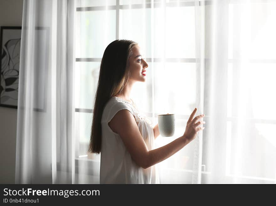 Young woman standing near window with curtains at home