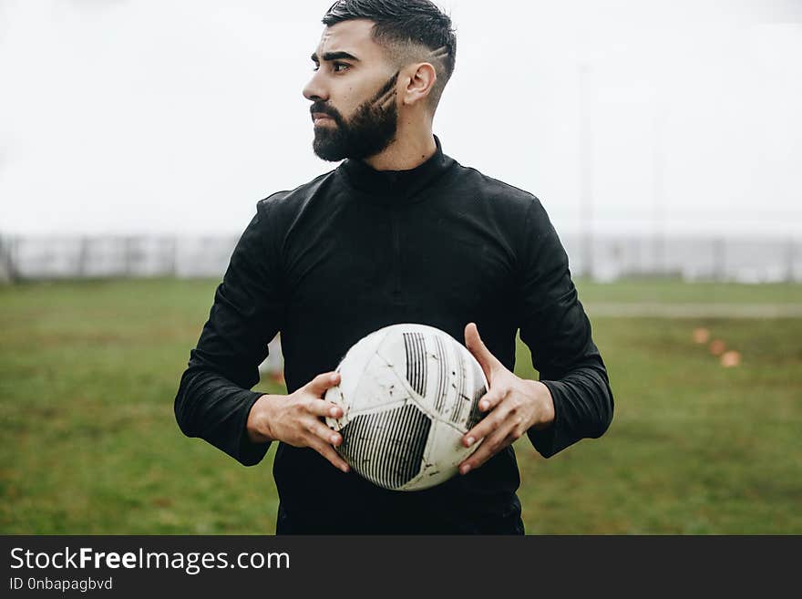 Portrait of a football player standing on field holding a ball looking away. Soccer player standing on ground holding a football during practice.