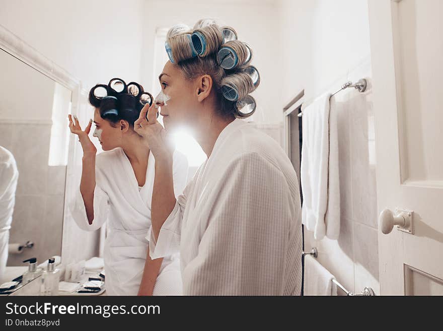 e view of two women in bathrobes applying cream on face standing in front of mirror. Women with curly rollers pinned on hair grooming at home.