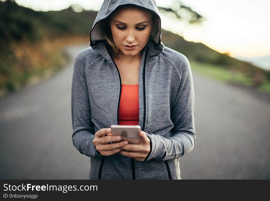 Fitness woman walking on street wearing a hooded sweat shirt looking at her mobile phone. Woman using her cell phone walking on an empty road.