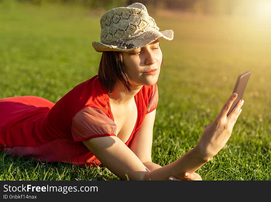 Attractive girl in red dress and yellow hat is lying on green grass and palying with her phone, toned image