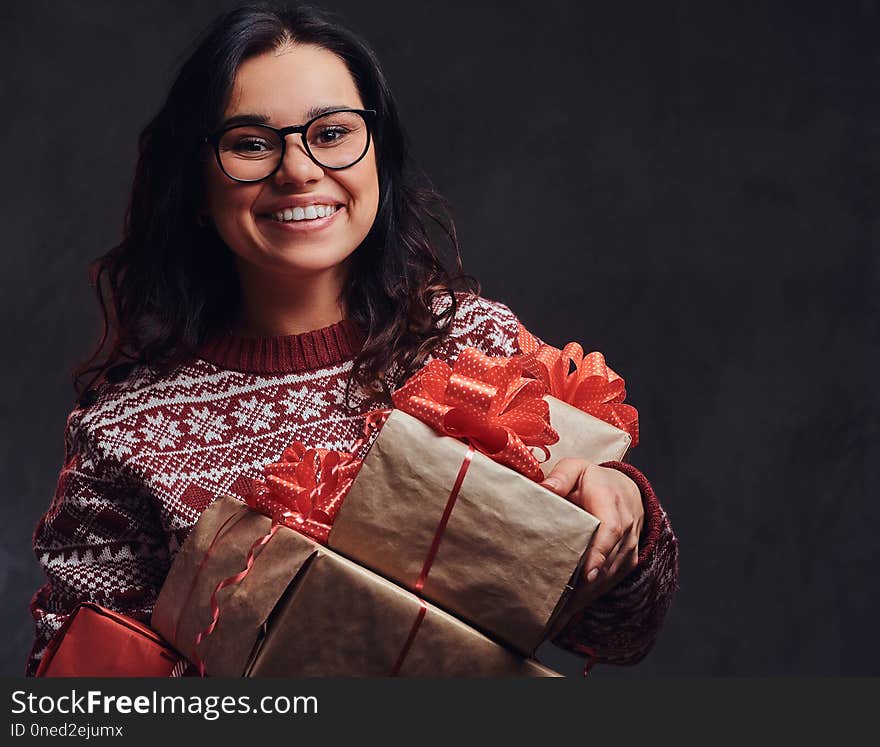 Christmas, Valentine`s Day, New year. Portrait of a happy brunette girl wearing eyeglasses and warm sweater holding a gifts boxes, isolated on a dark textured background.