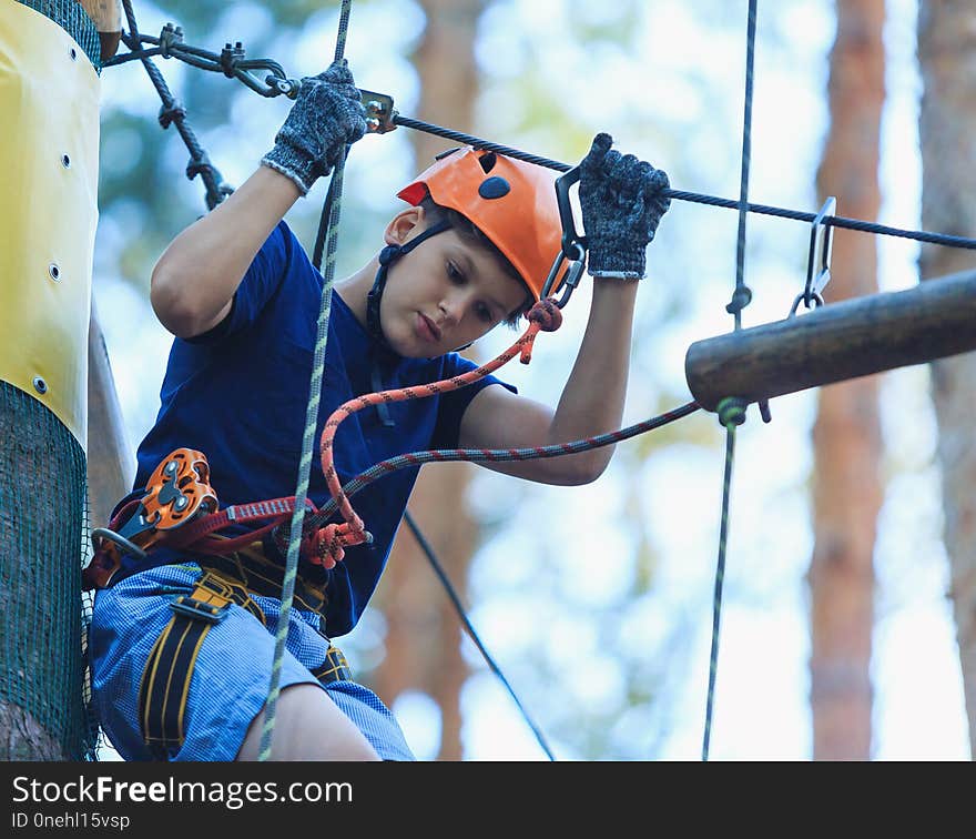 Child in forest adventure park. Kid in orange helmet and blue t shirt climbs on high rope trail. Agility skills and climbing outdoor amusement center for children. young boy plays outdoors.