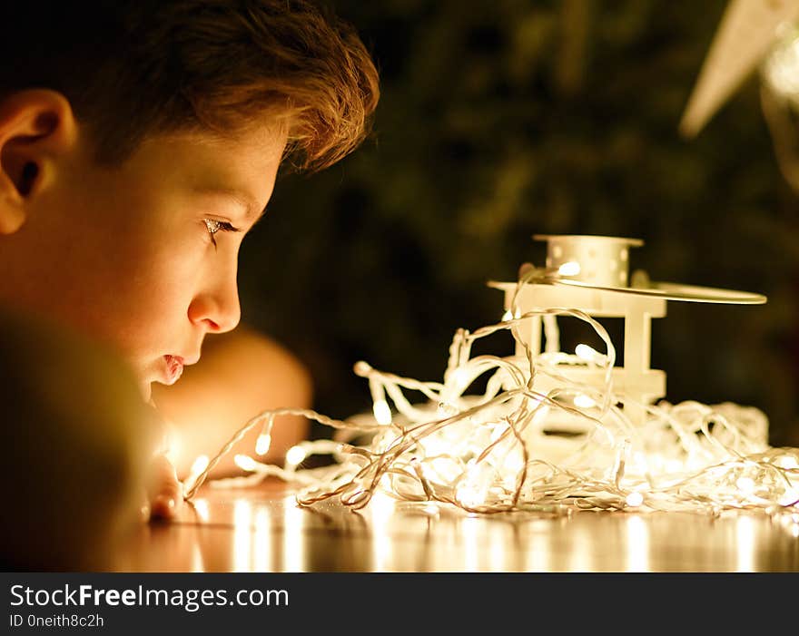 Cute young boy in white t shirt looks at lights in the evening at home in front of fir tree with lights. Winter holidays, New year and Christmas celebration concept.