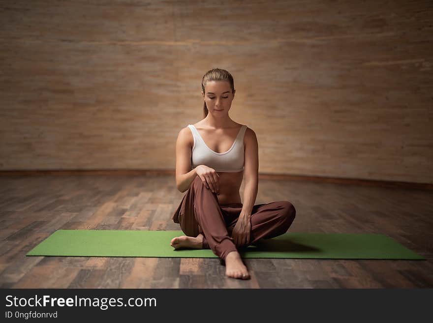 Peaceful young woman in comfortable clothes feeling relaxed while sitting on the yoga mat in empty room. Peaceful young woman in comfortable clothes feeling relaxed while sitting on the yoga mat in empty room