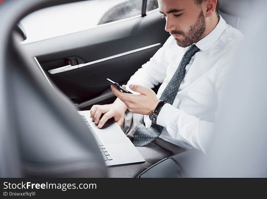 A young businessman working on laptop and talking on the phone while sitting in the car`s back. Works in motion