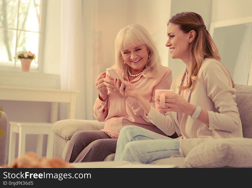 New smartphone. Happy cheerful women using a modern smartphone while sitting together on the sofa. New smartphone. Happy cheerful women using a modern smartphone while sitting together on the sofa