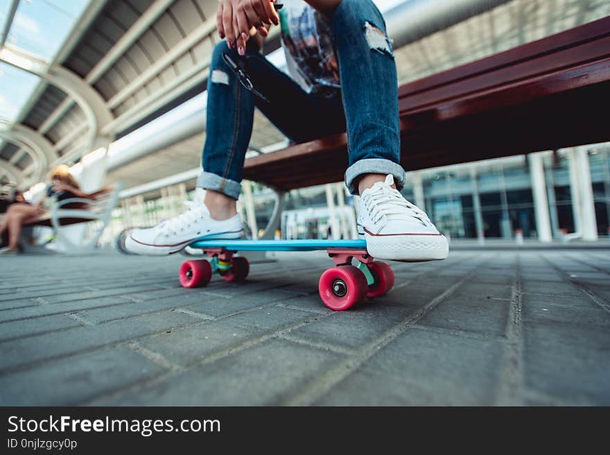 Girl sits on a bench in white sneakers and jeans, Close up of girls feet on a colourfull skateboard. Girl sits on a bench in white sneakers and jeans, Close up of girls feet on a colourfull skateboard.