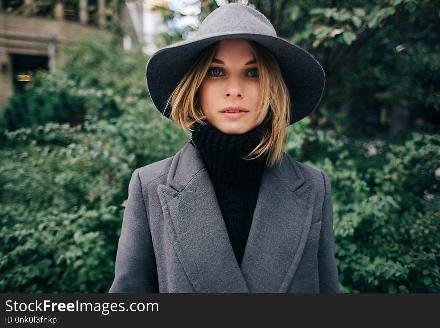 Photo of young blonde in gray coat on blurred background of city, green trees